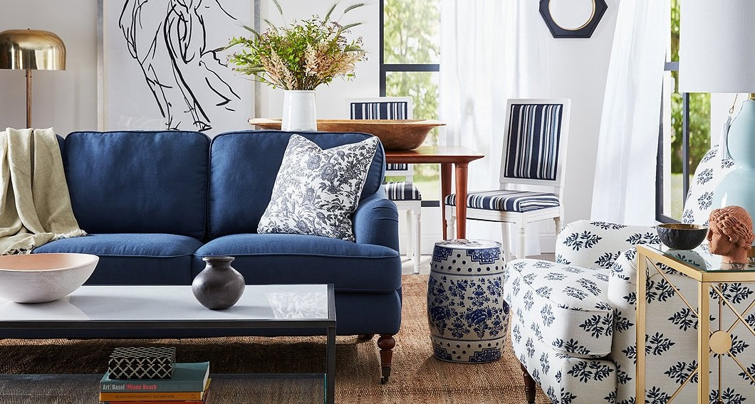 A bright living room features a blue sofa with a patterned pillow, a white and blue accent chair, and a table with blue and white striped chairs. There are plants, decorative vases, books, and a modern line-art piece on the wall with large windows in the background.