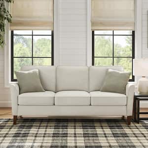 A dark grey Lifestyle Solutions Wesley 80.3" 3-seater sofa is placed on a plaid rug before two large windows with beige shades. A side table with a lamp sits to the right, and trees can be seen outside the windows.
