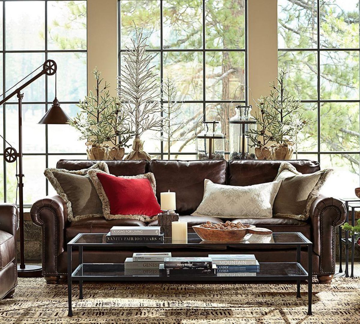 A cozy living room with a brown leather sofa cushioned with beige and one red pillow. Behind the couch, a wall of large windows reveals a scenic view of trees. A coffee table in front of the sofa holds candles, books, and a bowl. A floor lamp stands to the left.