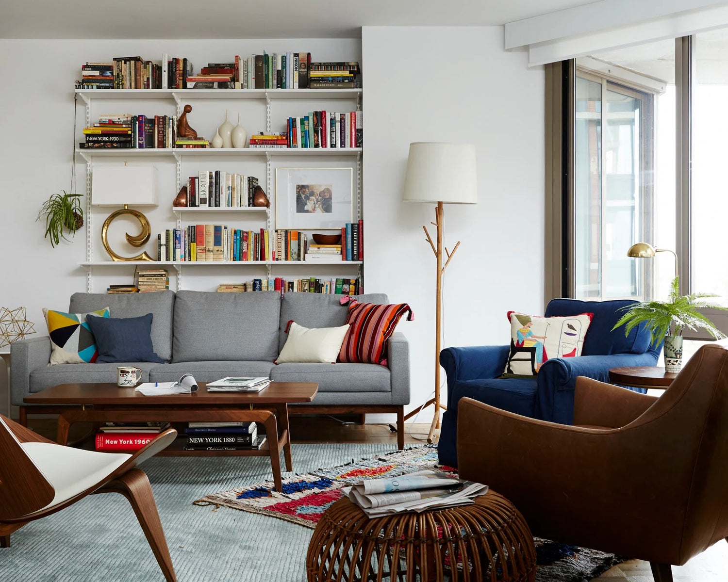 A modern living room features a gray sofa with colorful pillows, a blue armchair, and a wooden armchair. A tall floor lamp stands nearby. The back wall has floor-to-ceiling shelves filled with books and decor. Large windows bring in natural light, and a patterned rug lies on the floor.