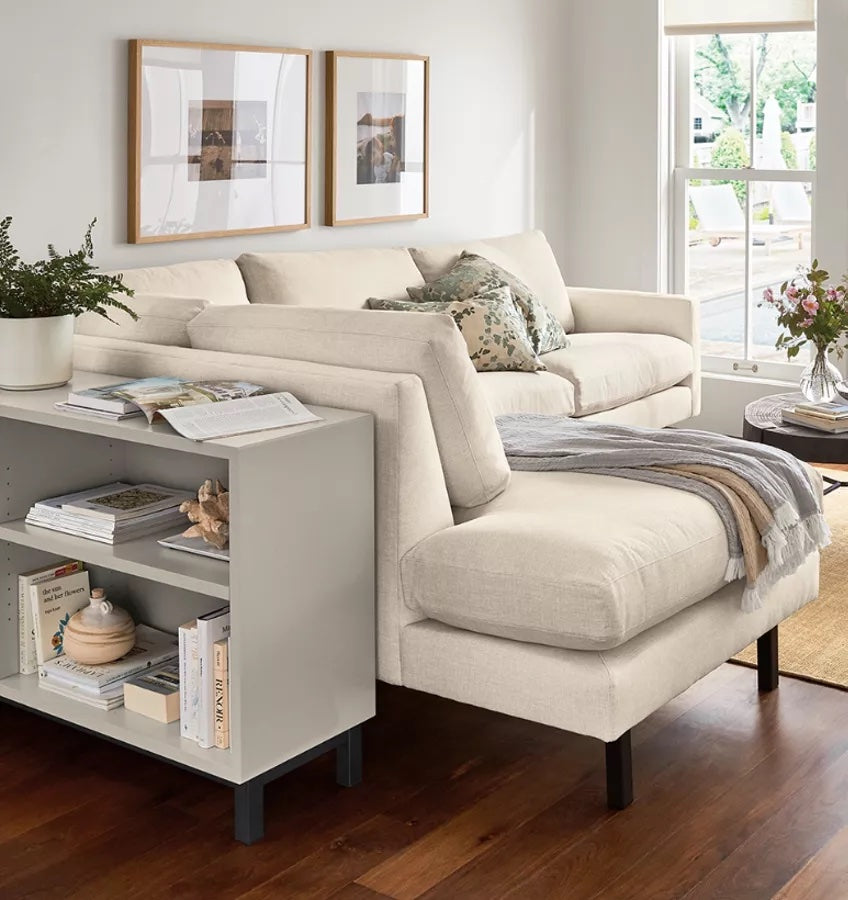 A cozy living room with a white L-shaped sofa adorned with patterned cushions and a gray throw blanket. Behind the sofa is a small white shelf holding books and decor. Two framed artworks hang on the wall above, and large windows let in natural light. A small round table with flowers sits to the right.