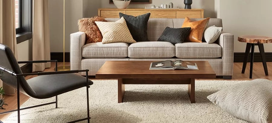 A cozy living room with a light gray sofa adorned with various patterned and textured throw pillows. In front of the sofa, there's a wooden coffee table with an open book. A black chair sits to the left, and a light rug covers the wooden floor.