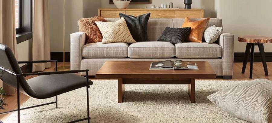 A cozy living room with a light gray sofa adorned with various patterned and textured throw pillows. In front of the sofa, there's a wooden coffee table with an open book. A black chair sits to the left, and a light rug covers the wooden floor.