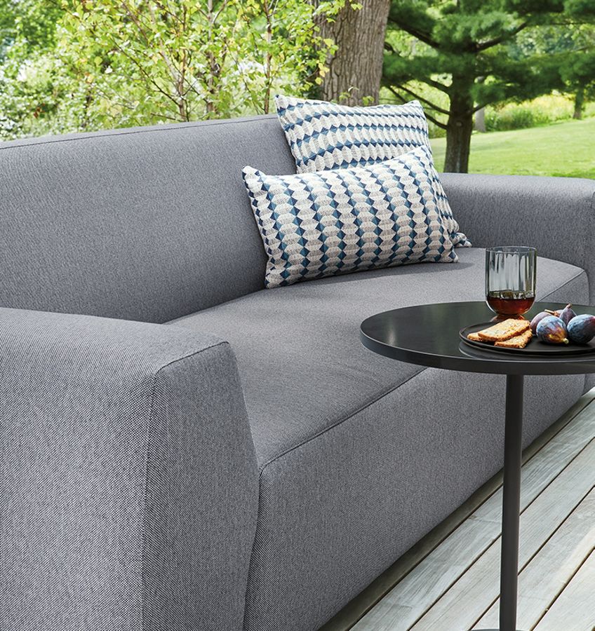 A modern gray outdoor sofa with two patterned cushions, situated on a wooden deck surrounded by lush greenery. A small black round table in front holds a glass of red beverage, a plate with snacks, and a few figs.