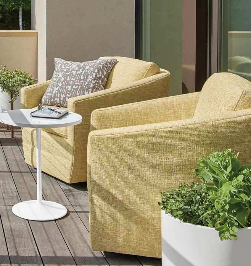 Two light yellow armchairs sit on a wooden deck outside. A small round white side table is placed between them, holding a book and a pair of glasses. Green plants in white pots decorate the space, with part of a sliding glass door visible in the background.