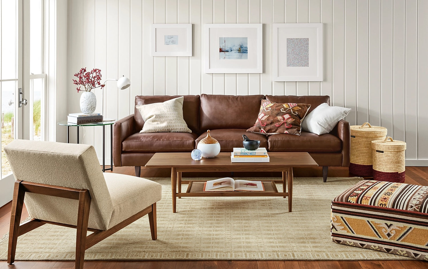 A modern living room with a brown leather sofa decorated with patterned cushions, a light beige armchair, and a wooden coffee table on a light patterned rug. The walls are adorned with framed artwork, and a small side table holds a vase with red flowers.