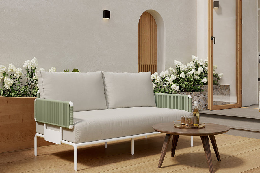 A serene patio scene showcases the Sylva Outdoor Modular Sofa by Acanva, featuring modern design with light green armrests and white cushions. In front stands a round wooden coffee table adorned with drinks and glasses. White flowers and lush greenery fill a wooden planter by a pale wall with an arched doorway.