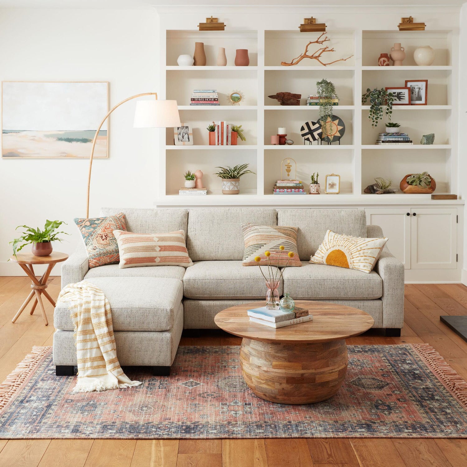 A cozy, modern living room with a light gray sectional sofa adorned with patterned throw pillows. A round wooden coffee table sits on a vintage rug. The wall behind features white built-in shelves with various plants, books, and decor items, including a painting and a lamp.