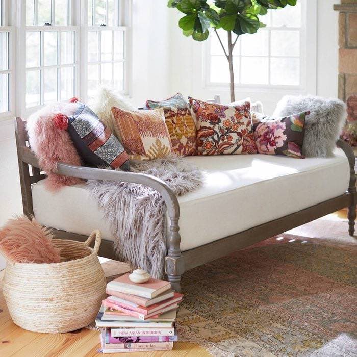 A cozy daybed with white cushions is adorned with a variety of colorful pillows and gray faux fur throws. A woven basket containing more pillows is placed nearby, and a stack of books sits on the floor. A leafy green plant and large windows brighten the space.