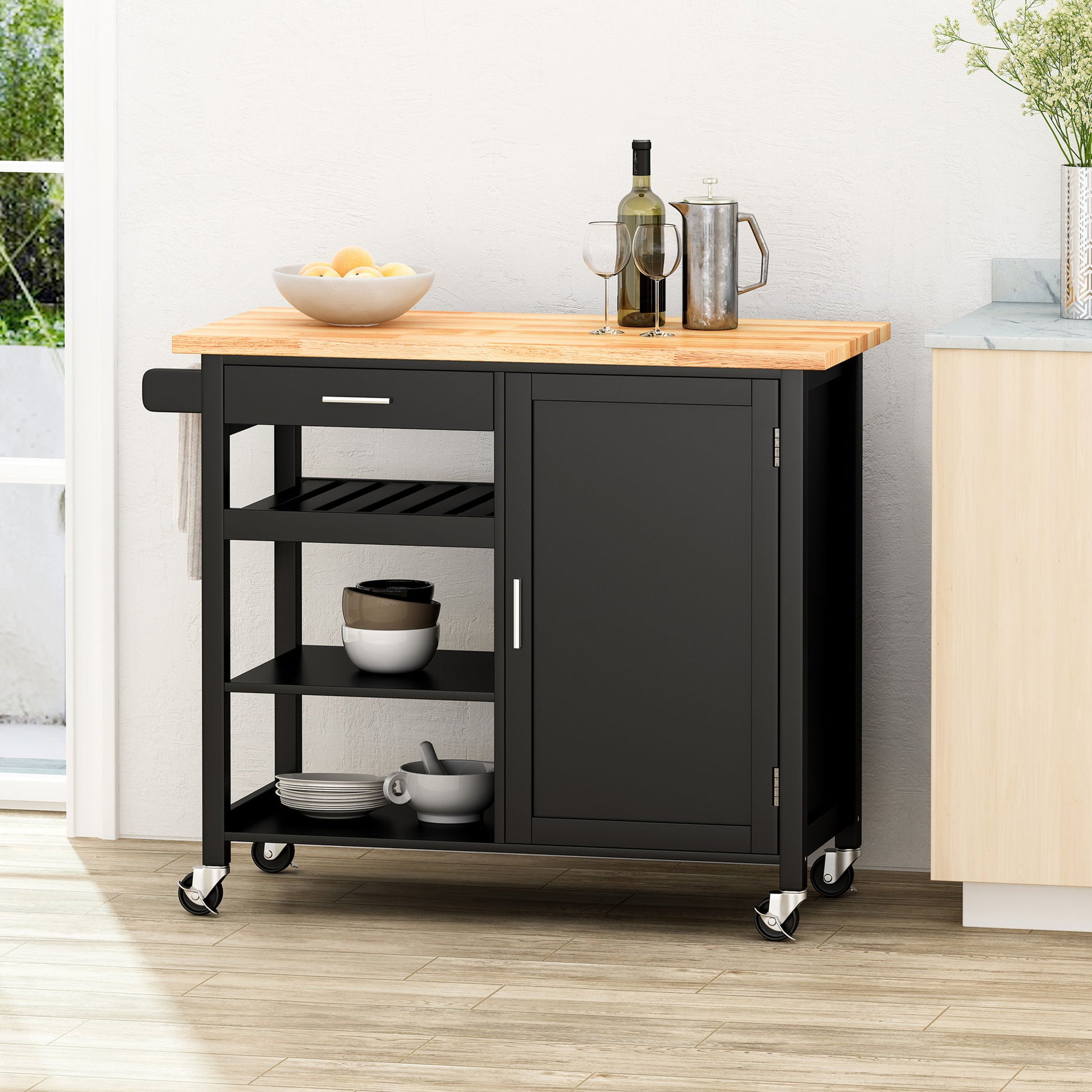 The Contemporary Design Kitchen Cart With Wheels, featuring a black finish and wooden top, holds lemons, wine glasses, a wine bottle, and a pitcher. It offers open shelves for dishes, a closed cabinet, and sits on wheels in a bright kitchen.