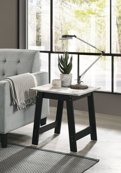 A modern living room features a gray sofa, striped throw blanket, and the Kenzo End Table with Faux Marble Top Finish in black, which holds two potted plants, a lamp, a candle, and decor; large windows are in the background.