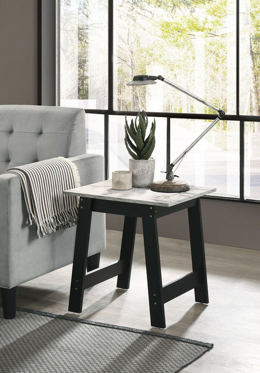 A modern living room features a gray sofa, striped throw blanket, and the Kenzo End Table with Faux Marble Top Finish in black, which holds two potted plants, a lamp, a candle, and decor; large windows are in the background.
