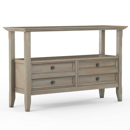 The Amherst Console Sofa Table is handcrafted from wood with a weathered gray finish, featuring an open top shelf, four drawers with round metal knobs, and stands on four tapered legs.