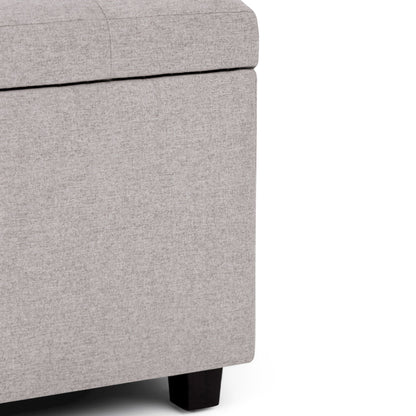 Close-up of the Cosmopolitan - Upholstered Storage Ottoman in light gray, featuring dark wooden legs. Shown against a white background, only part of the lid and two sides are visible.
