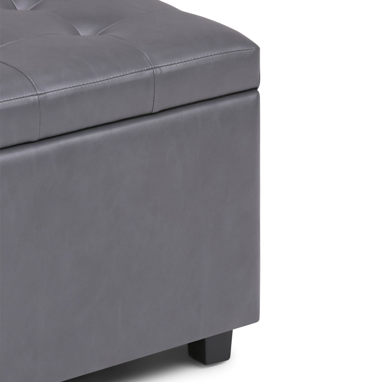 Close-up of the corner of the Cosmopolitan - Upholstered Storage Ottoman in gray, featuring tufted detailing on the top, a smooth padded surface, and short black legs.