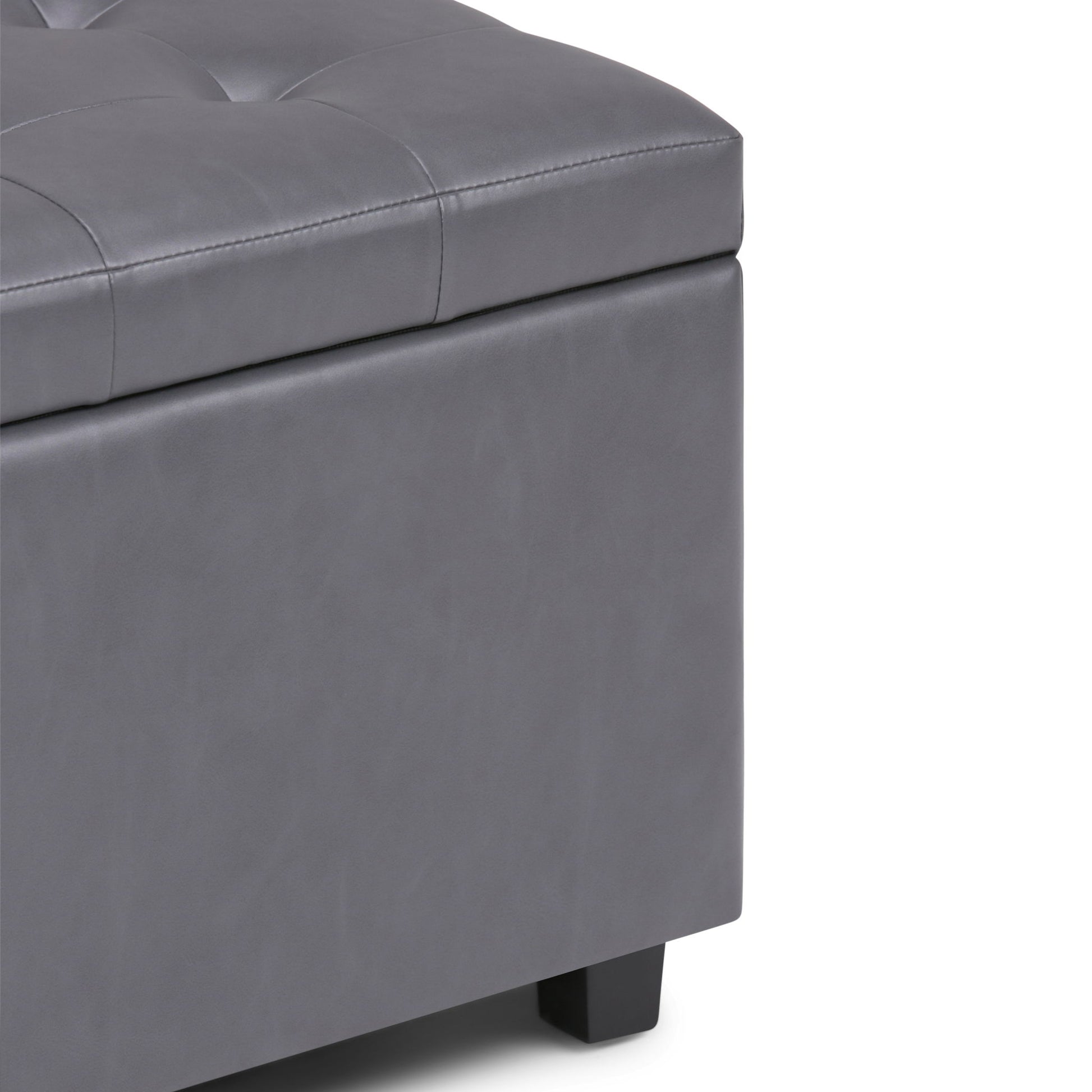 Close-up of the corner of the Cosmopolitan - Upholstered Storage Ottoman in gray, featuring tufted detailing on the top, a smooth padded surface, and short black legs.