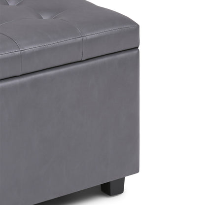 Close-up of the corner of the Cosmopolitan - Upholstered Storage Ottoman in gray, featuring tufted detailing on the top, a smooth padded surface, and short black legs.