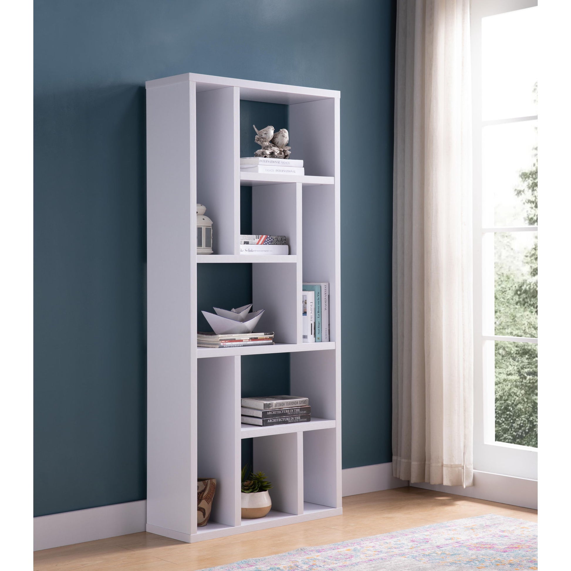 The Versatile Display Cabinet, a modern white piece with asymmetrical shelves, holds books, decor, and a plant. It stands against a teal wall by a window with sheer curtains on a light wood floor.