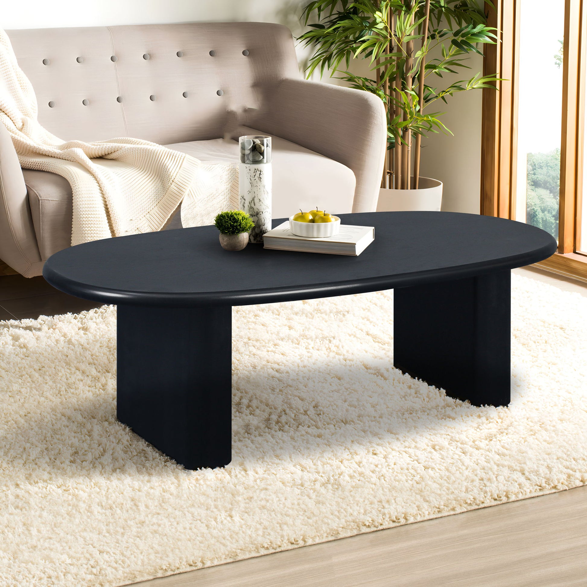 A modern living room features the Tapered Tabletop Coffee Table in Antique Black on a white fluffy rug, set before a beige sofa with a blanket. Large windows let in sunlight, a plant sits nearby, and books and decor items adorn the table.