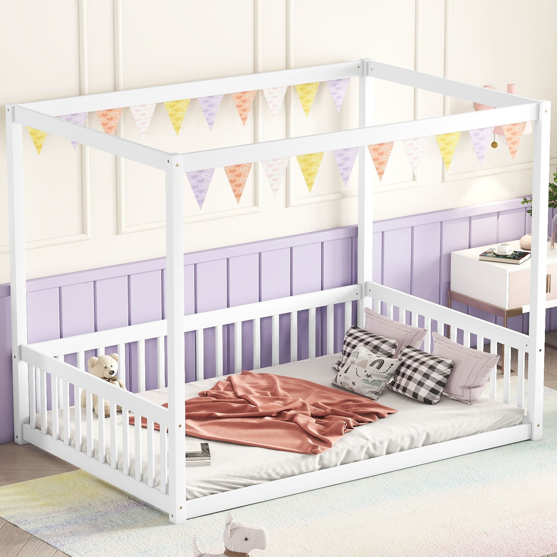 A Canopy Frame Floor Bed With Fence, Guardrails sits in a pastel room with a pink blanket, plaid pillows, and a teddy bear. Multicolored triangle bunting hangs above, and a white nightstand with toys is visible in the background.