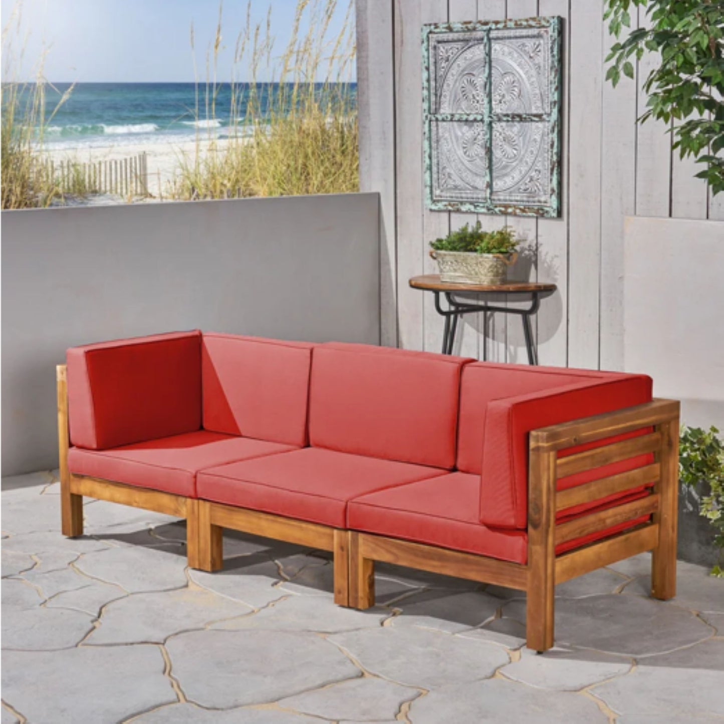 The Oana - 3 Piece Sofa Set With Water Resistant Cushions, featuring red cushions, is arranged on a stone patio beside a small table with plants and set against a gray fence, with sand dunes and the ocean visible in the background.