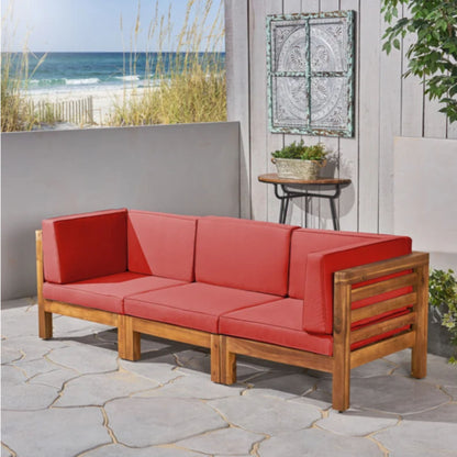 The Oana - 3 Piece Sofa Set With Water Resistant Cushions, featuring red cushions, is arranged on a stone patio beside a small table with plants and set against a gray fence, with sand dunes and the ocean visible in the background.