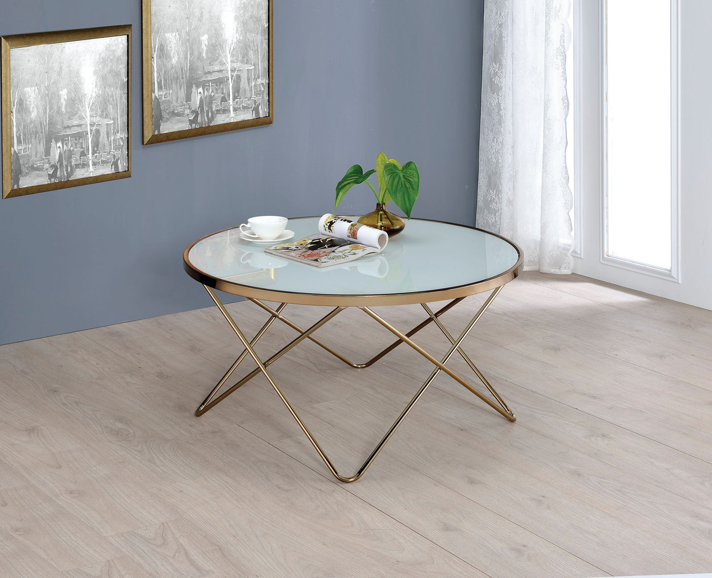 The Valora - Glass Coffee Table, featuring a gold metal frame, sits on a light wood floor. On it are a cup and saucer, a rolled magazine, and a small potted plant. Two framed artworks hang on the nearby gray wall.