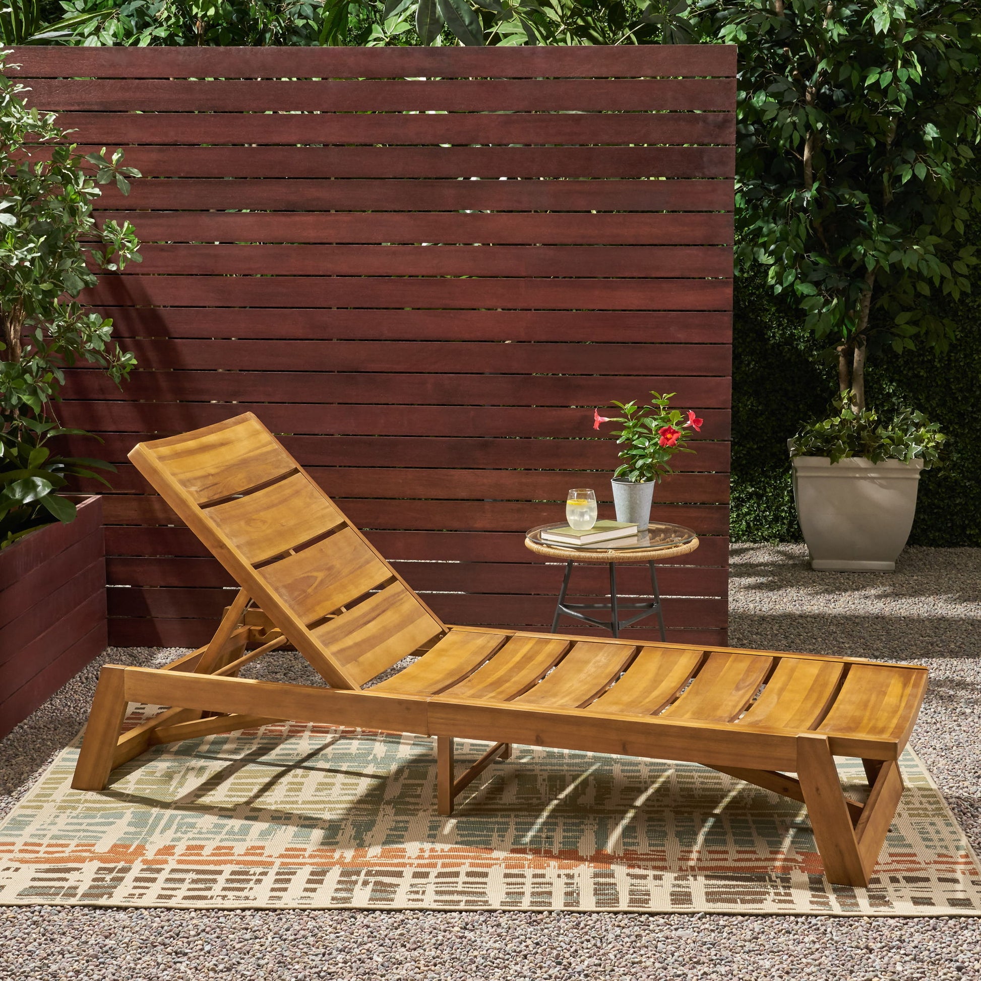 An Outdoor Chaise Lounge With Adjustable Seating And Slat Panel in yellow brown sits on a patterned rug beside a small round table with a drink and potted flower, all set against a wooden fence with lush green plants behind.