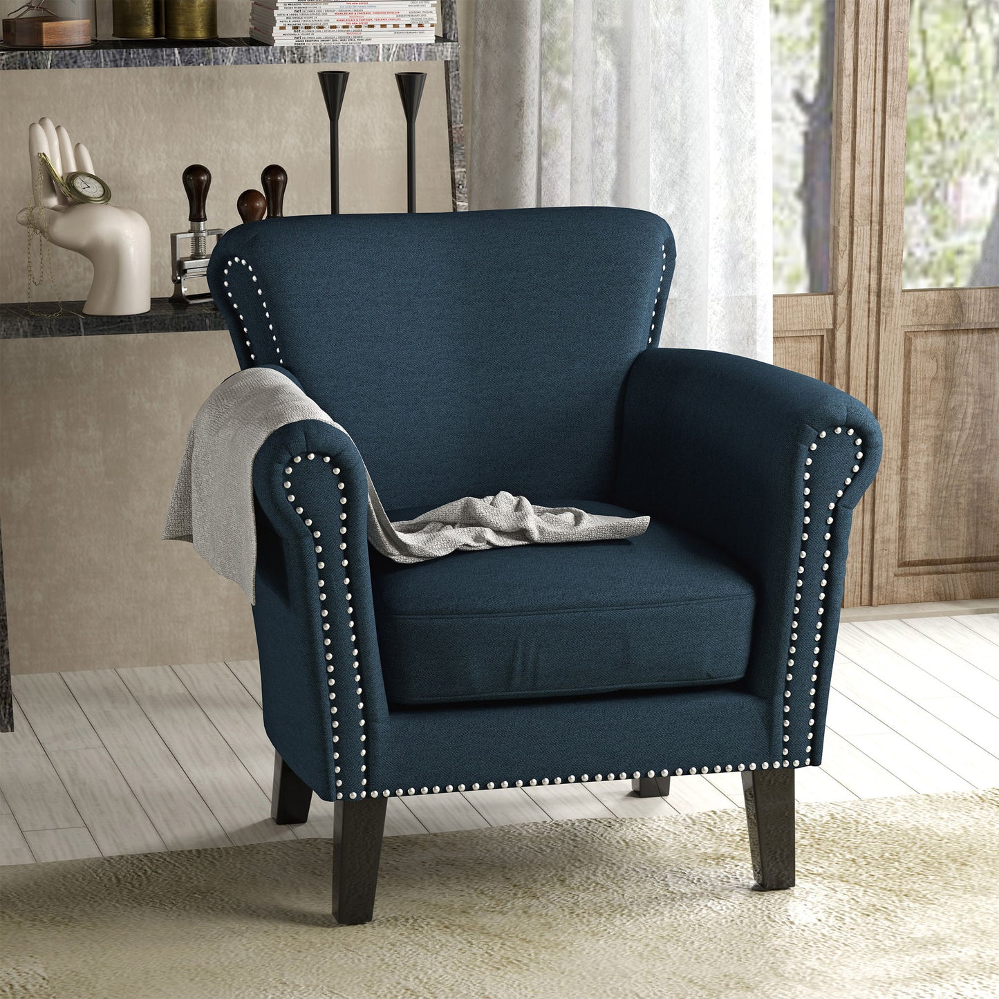 The Elegant Club Chair With Nailhead Trim And Birch Wood Legs, upholstered in dark blue, sits on a light floor. A gray blanket is draped over one armrest, with shelves and decorative items visible in the background.