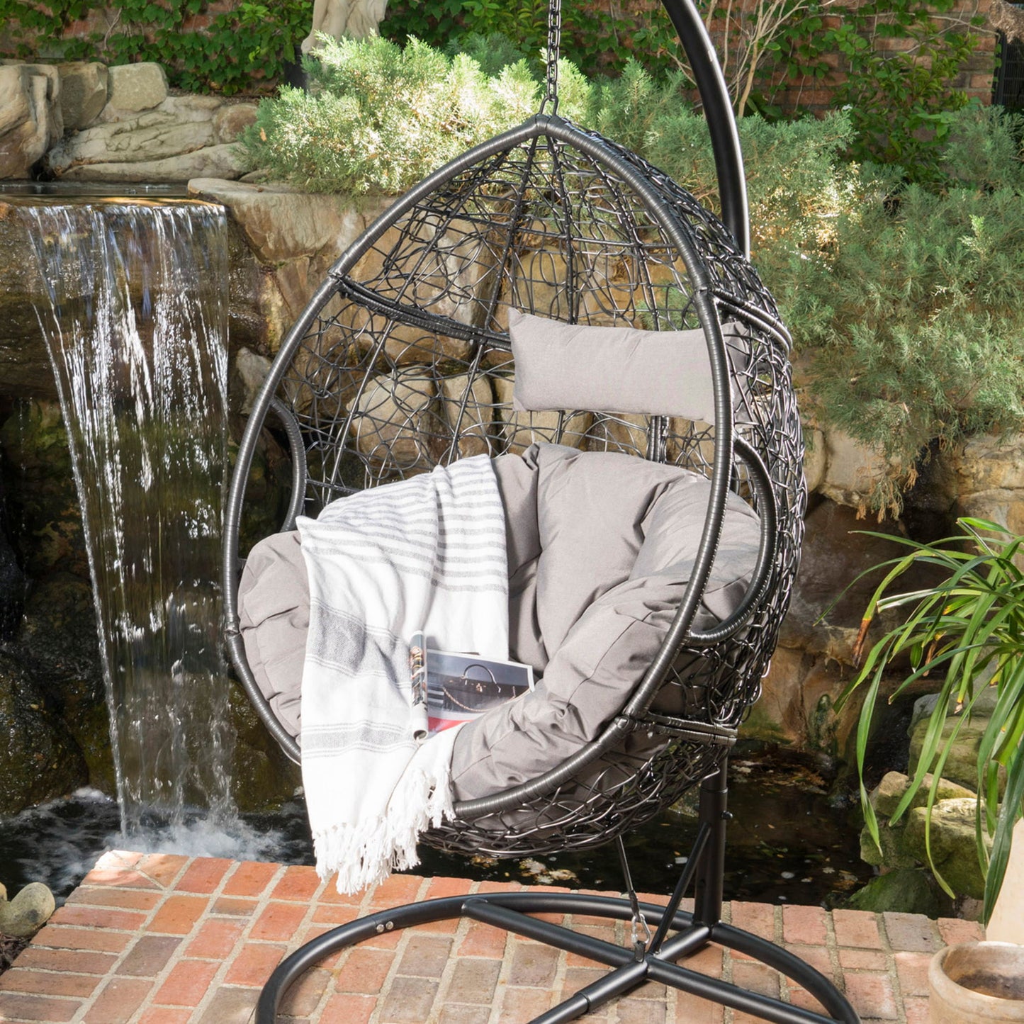 The Kylie Boho Style Hanging Basket Chair Rattan with water-resistant gray cushions sits on a brick patio by a pond with a waterfall, surrounded by lush greenery and accented with a striped blanket.