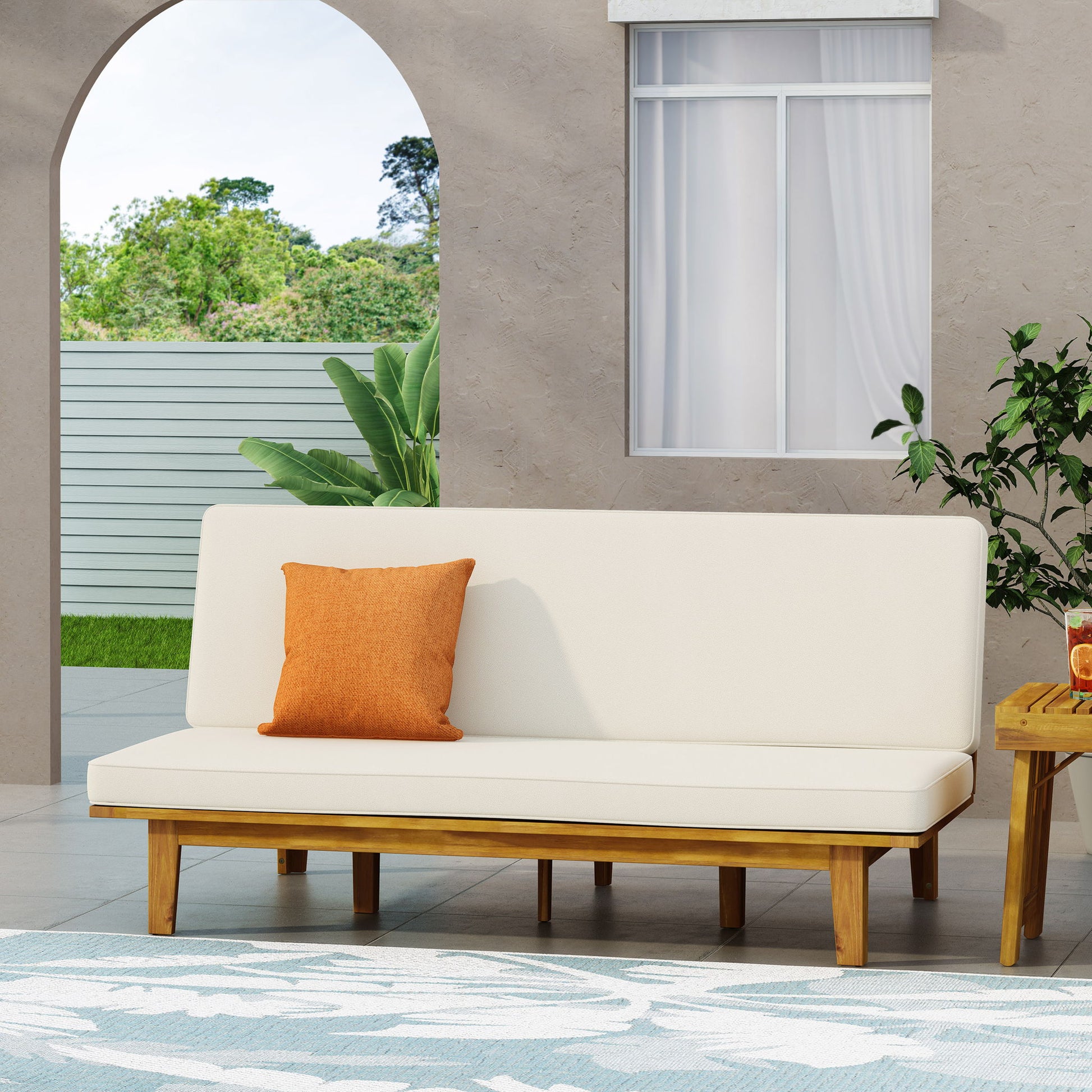 A modern patio showcases the Outdoor Elegance Daybed with a convertible beige cushion frame, accented by an orange pillow. A small wooden table with drinks sits nearby, while lush greenery and a fence are visible through an arched opening.
