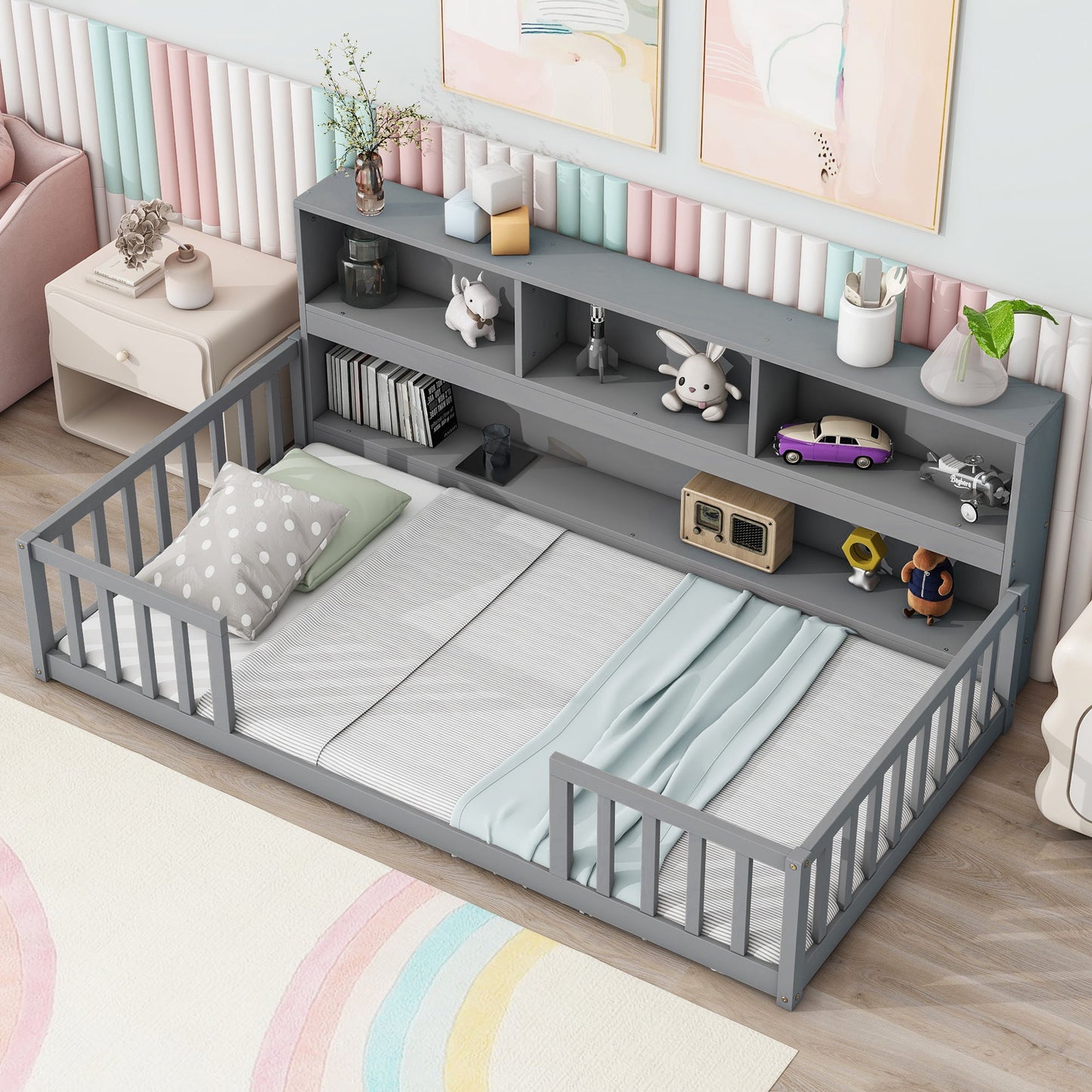 The Floor Bed With Bedside Bookcase features gray guardrails, a headboard with shelves for toys and books, and comes with pillows and a blanket in a pastel-themed room with striped wall panels and a nearby side table.