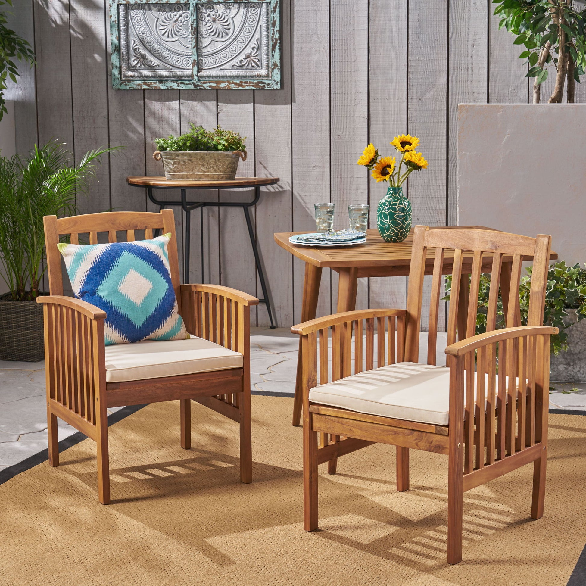 Two Casa - Acacia Dining Chairs with white cushions and a blue pillow are placed by a wooden table holding sunflowers, glasses, and plates. The scene is an outdoor patio decorated with plants and wall art.