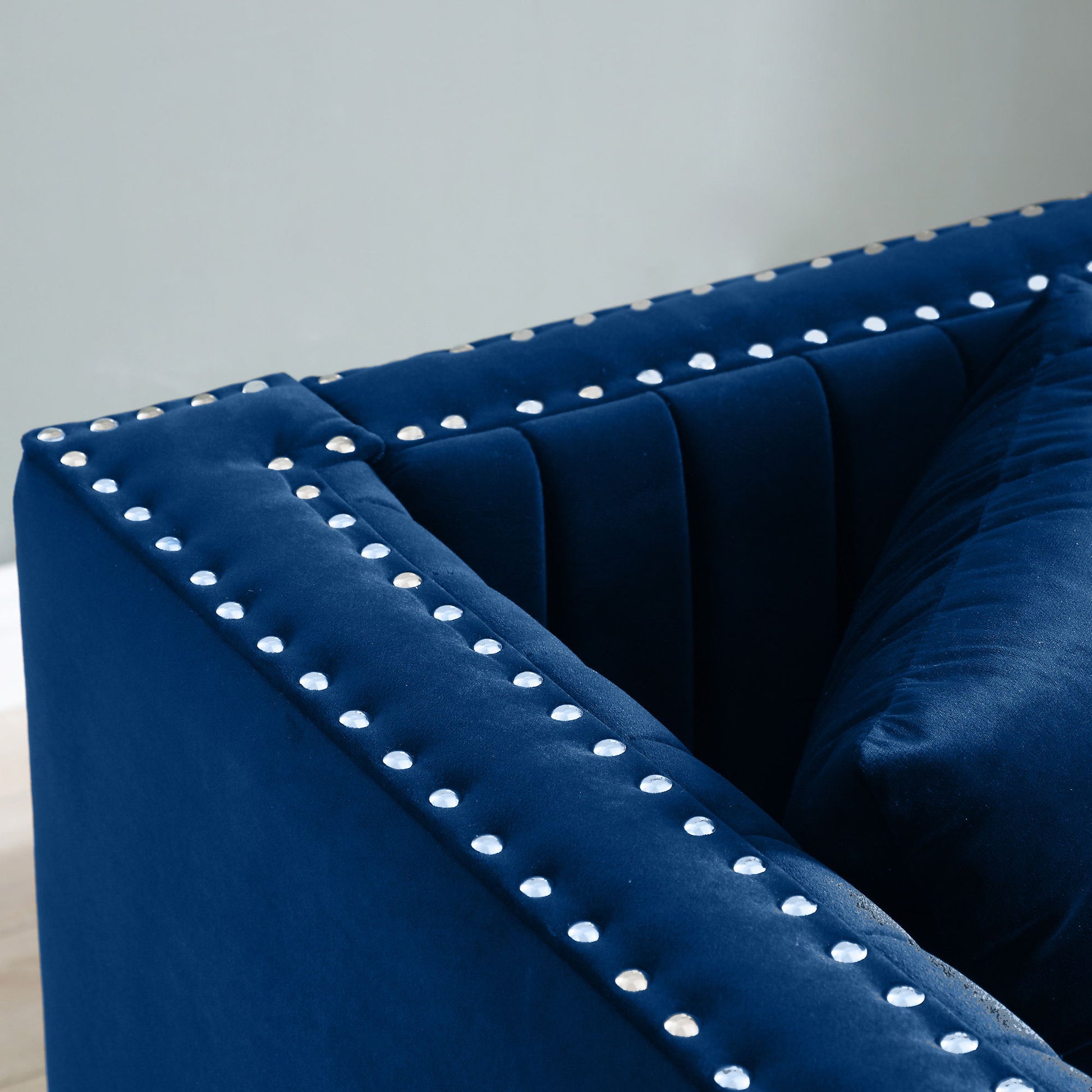 Close-up of the Contemporary Vertical Channel Tufted Sofa, a navy blue velvet modern upholstered couch with silver studded accents on the arms and backrest, shown with 2 pillows against a light gray wall.
