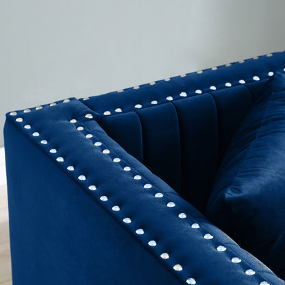 Close-up of the Contemporary Vertical Channel Tufted Sofa, a navy blue velvet modern upholstered couch with silver studded accents on the arms and backrest, shown with 2 pillows against a light gray wall.