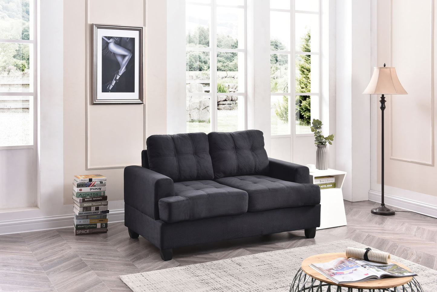 A modern living room with a Stylish Transitional Loveseat For Two in black, a stack of books, a round coffee table with magazines, a floor lamp, side table with plants, large windows, and a framed photo of a woman's legs on the wall.