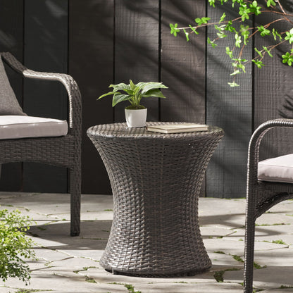 A Thrive Hourglass Rattan Table with no glass top holds a potted plant and two plates, paired with two cushioned wicker chairs on a stone patio, surrounded by greenery and a dark wooden fence.