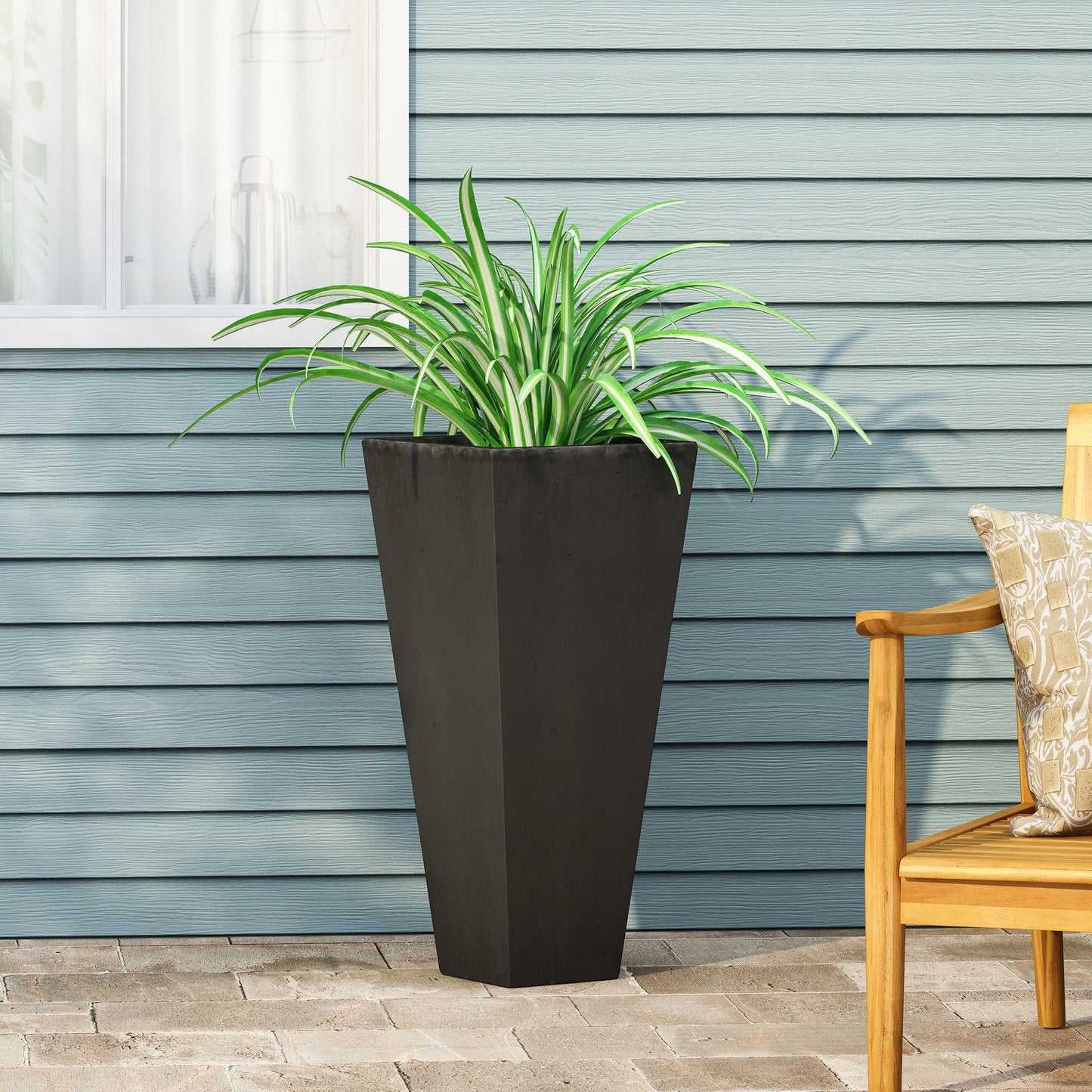 An Outdoor Modern Cast Stone Planter with a spider plant sits on a stone patio by a blue horizontal siding wall. To the right, part of a wooden chair with a patterned cushion is visible. A window appears in the background.