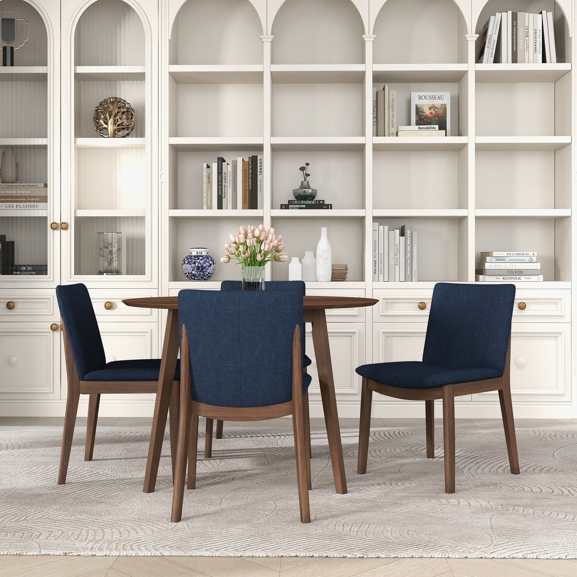 A round wooden dining table with a vase of pink tulips is surrounded by four Laura Mid-Century Modern Solid Wood Dining Chairs. A white built-in bookshelf filled with books and decor stands behind the table.