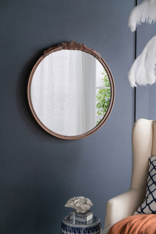 The Round Hand Carved Rose Antique Mirror Frame hangs on a dark gray wall, reflecting sheer white curtains and green leaves. Nearby are an armchair, pillow, blanket, and a side table topped with coral decor.
