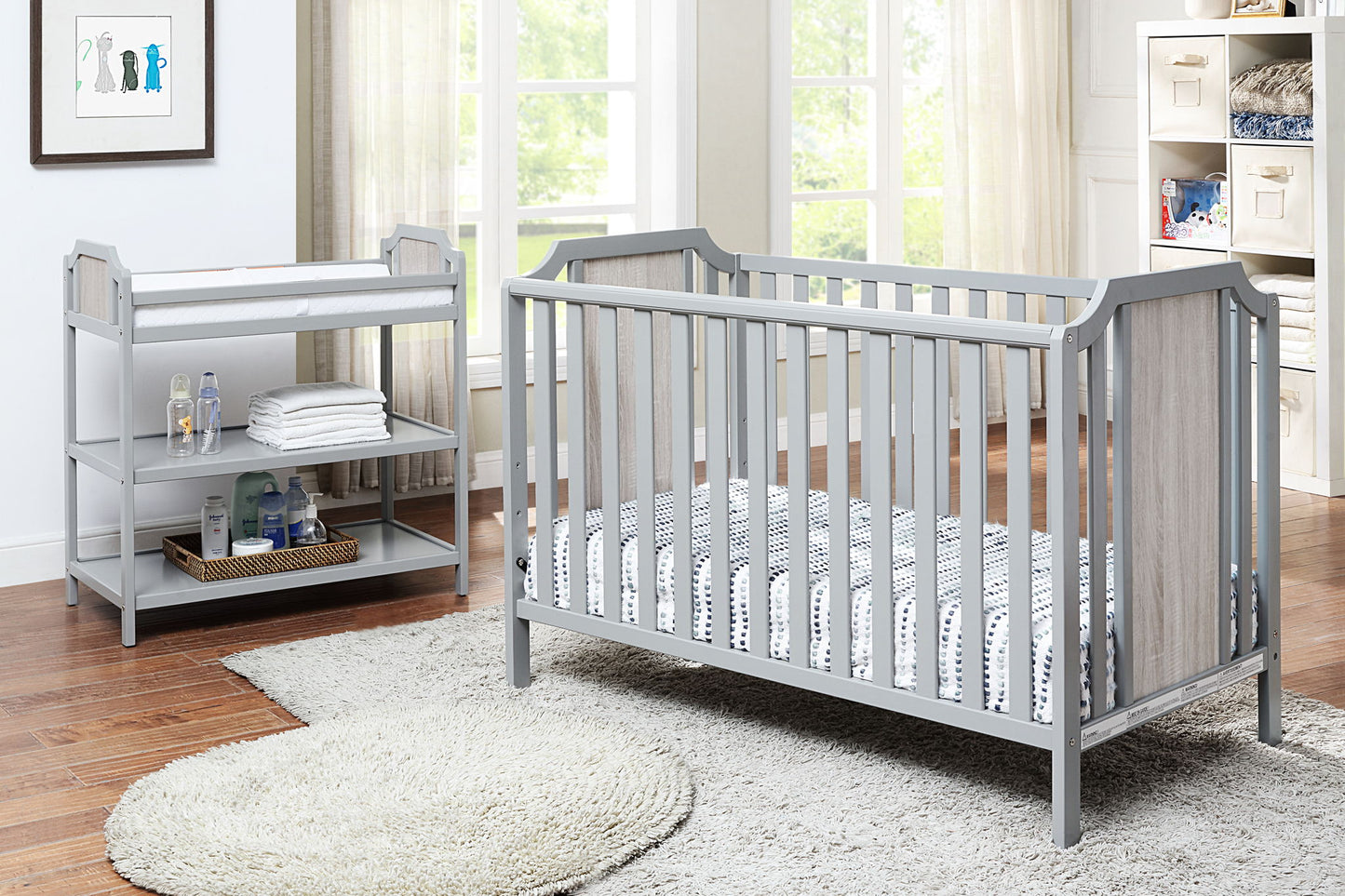 A bright nursery with a gray crib and mattress, a Brees - Changing Table with shelves for baby supplies, a round rug on hardwood floors, and large windows letting natural light in.