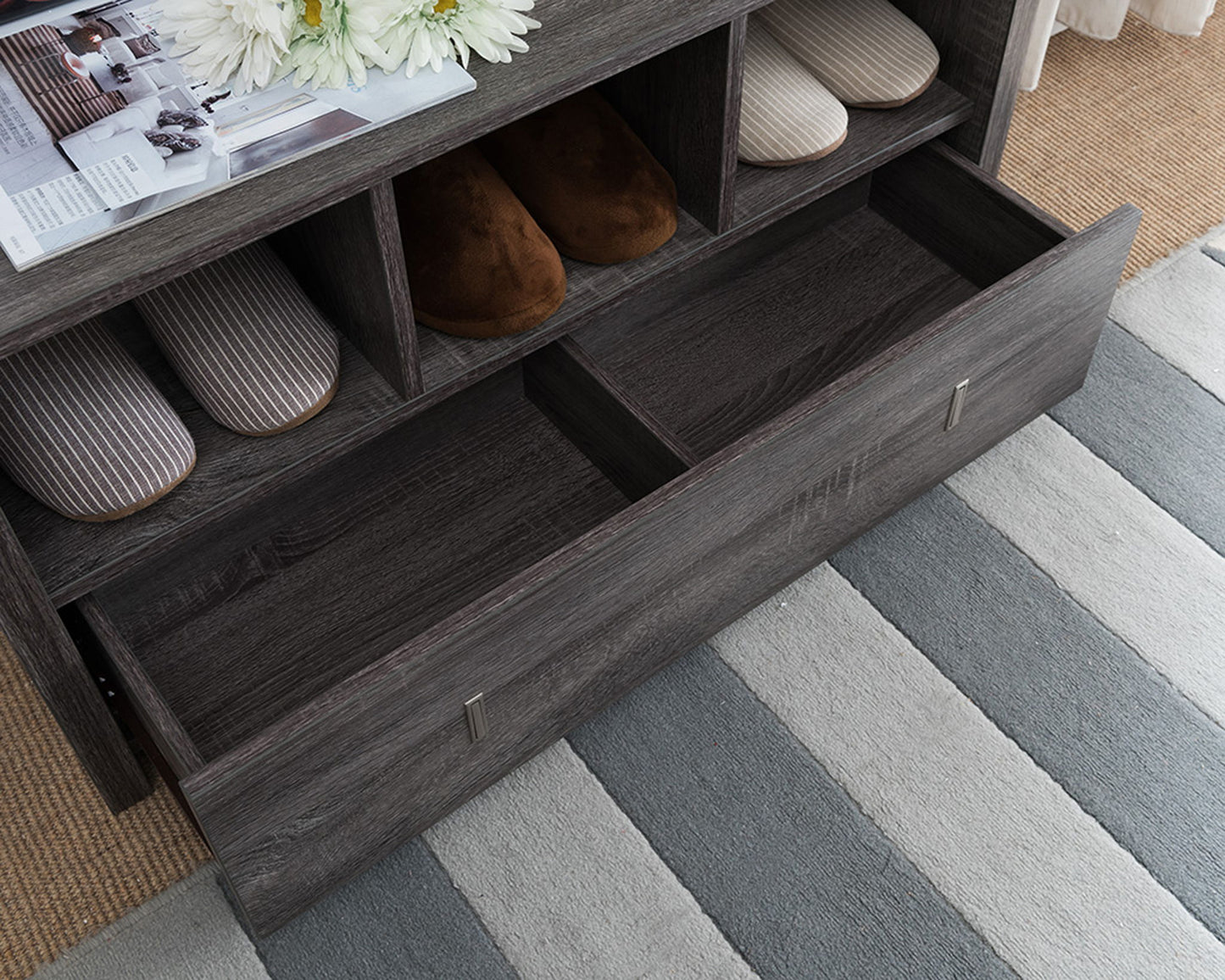 A Shoe Entry Bench With Three Shelves and a divided-compartment drawer holds striped and brown slippers, all set on a gray and white striped rug, with a magazine and flowers placed on top.
