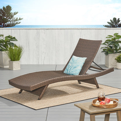 The Salem - Chaise Lounge with a blue leaf pillow rests on a patio rug amid potted plants. A small wooden table with drinks and fruit sits nearby, while a white fence frames an ocean view in the background.