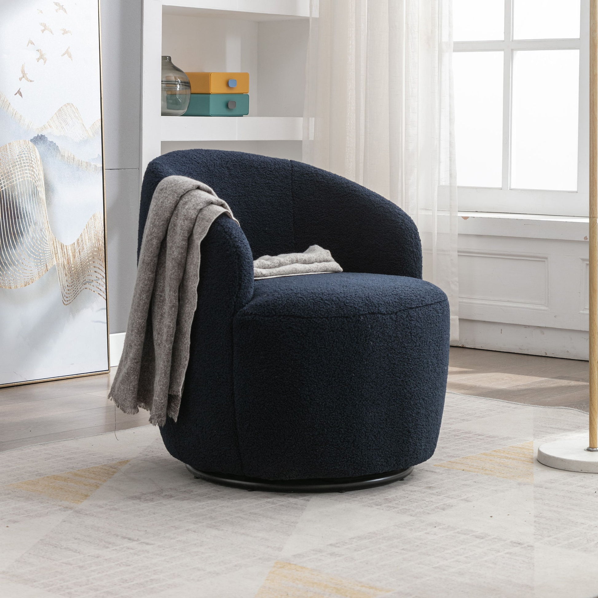 A Teddy Fabric Swivel Accent Armchair Barrel Chair with a powder-coated metal ring sits on a light patterned rug. Draped with a light gray blanket, it features a curved backrest. Shelves, sheer-curtained window, and abstract art complete the background.