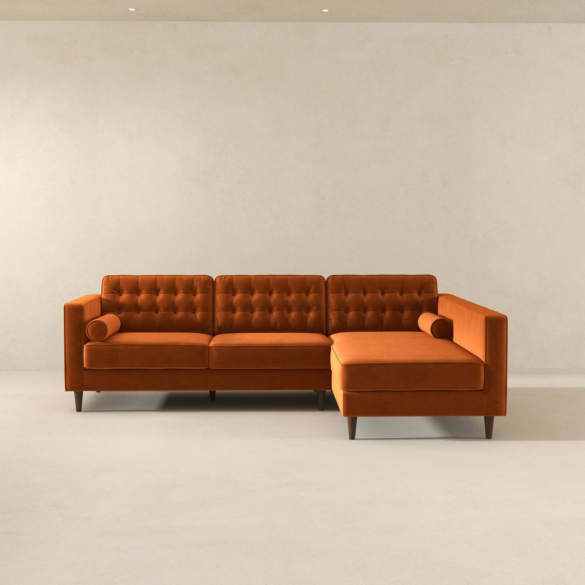 The Christian sectional sofa features mid-century modern design with orange tufted upholstery, two cylindrical pillows, and wooden legs, set in a minimal room with plain walls and a light floor.