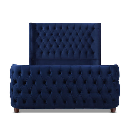 The Brooklyn Tufted Panel Bed features a navy blue velvet frame with high padded headboard and matching footboard, both showcasing diamond button tufting. It stands on short, dark wooden legs for a stylish finish.