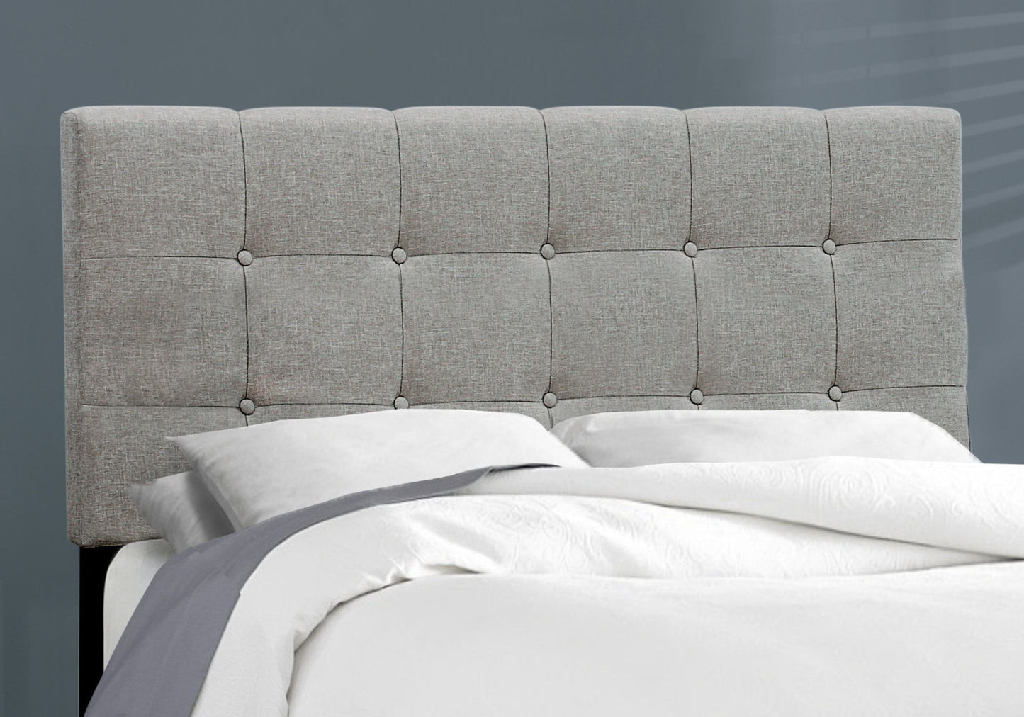 A neatly made bed with white sheets, a gray blanket, and a light gray transitional upholstered frame-only headboard stands against a plain gray wall.