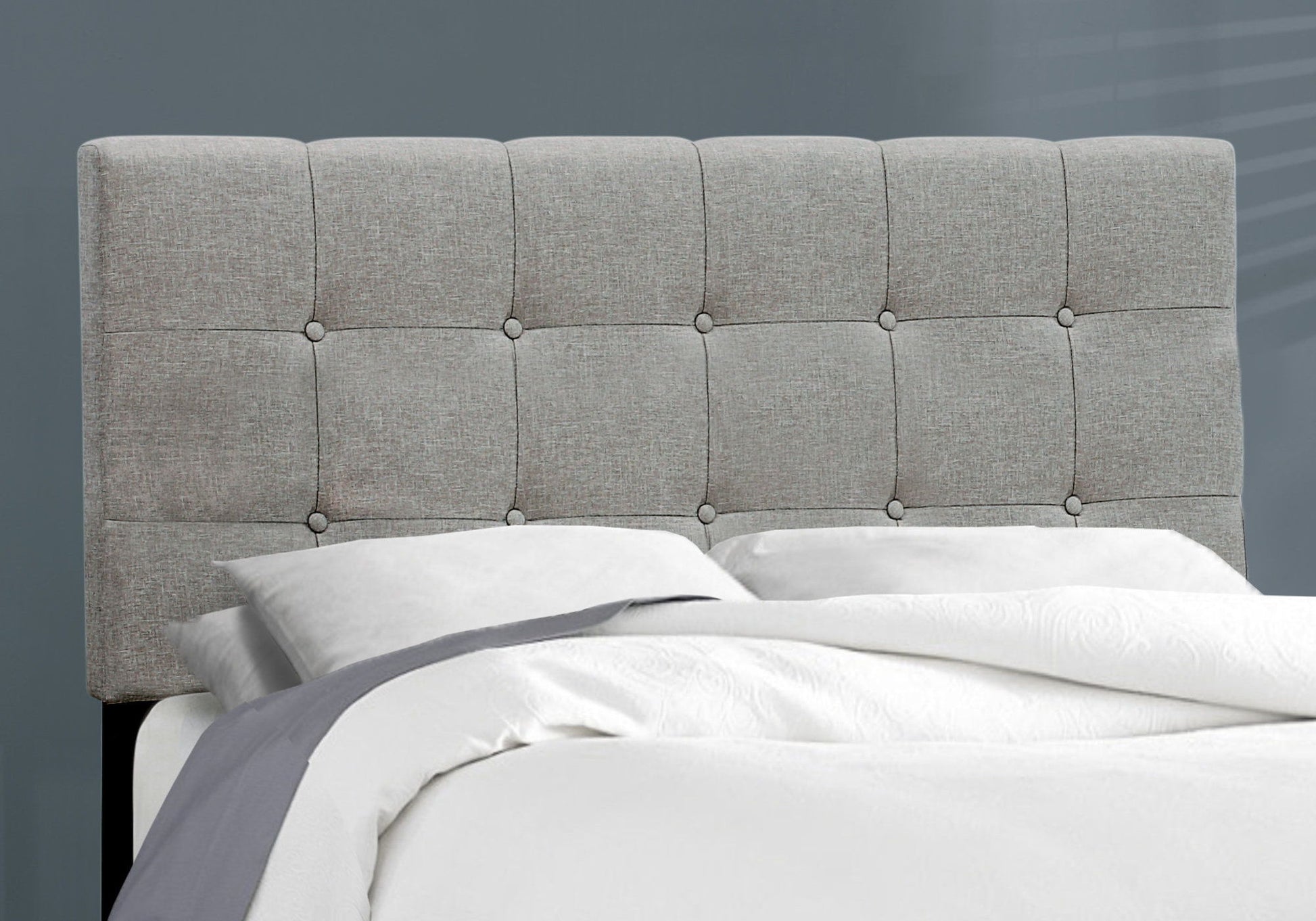 A neatly made bed with white sheets, a gray blanket, and a light gray transitional upholstered frame-only headboard stands against a plain gray wall.