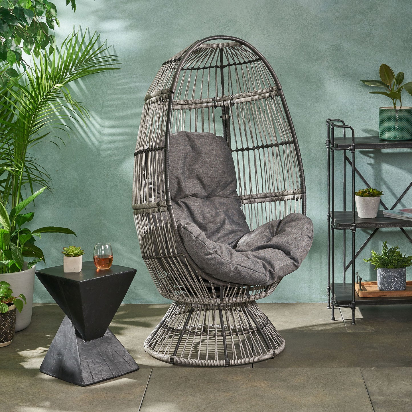 The Patio Swivel Basket Egg Chair With Cushion in gray sits beside a black geometric side table holding a drink and plant, surrounded by potted greenery against a light teal wall on the patio.