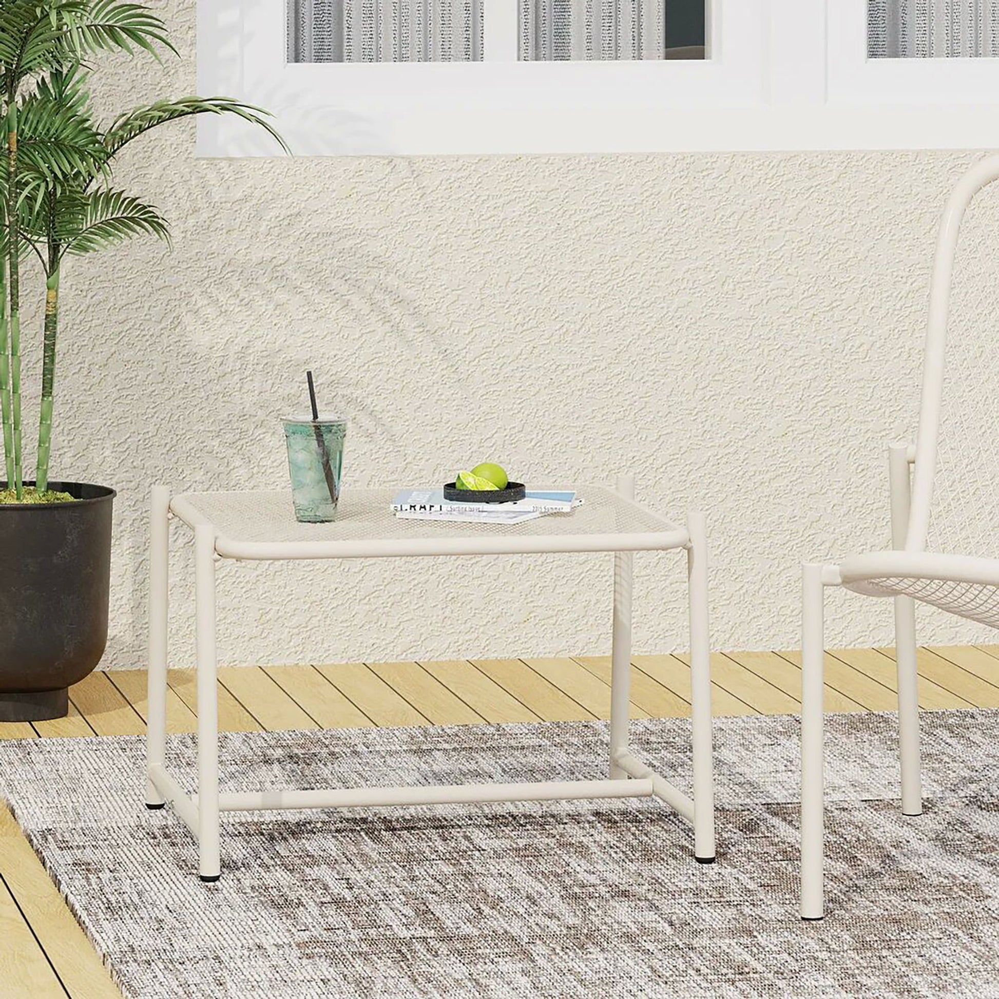The Conrad Side Table Outdoor, light-colored and compact, holds a drink and book on a rug beside a chair. A potted plant is nearby, with sheer-curtained windows in the background.